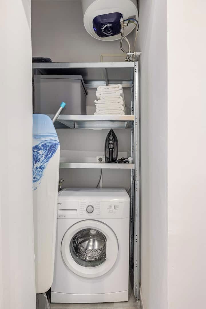 A compact laundry room with a washing machine, stacked towels, an iron, and a security camera.
