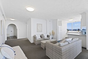 Ein modernes Wohnzimmer mit einer Couch, zwei Sesseln, einer Essecke mit einem Kühlschrank und Blick auf die Stadt durch große Fenster.
