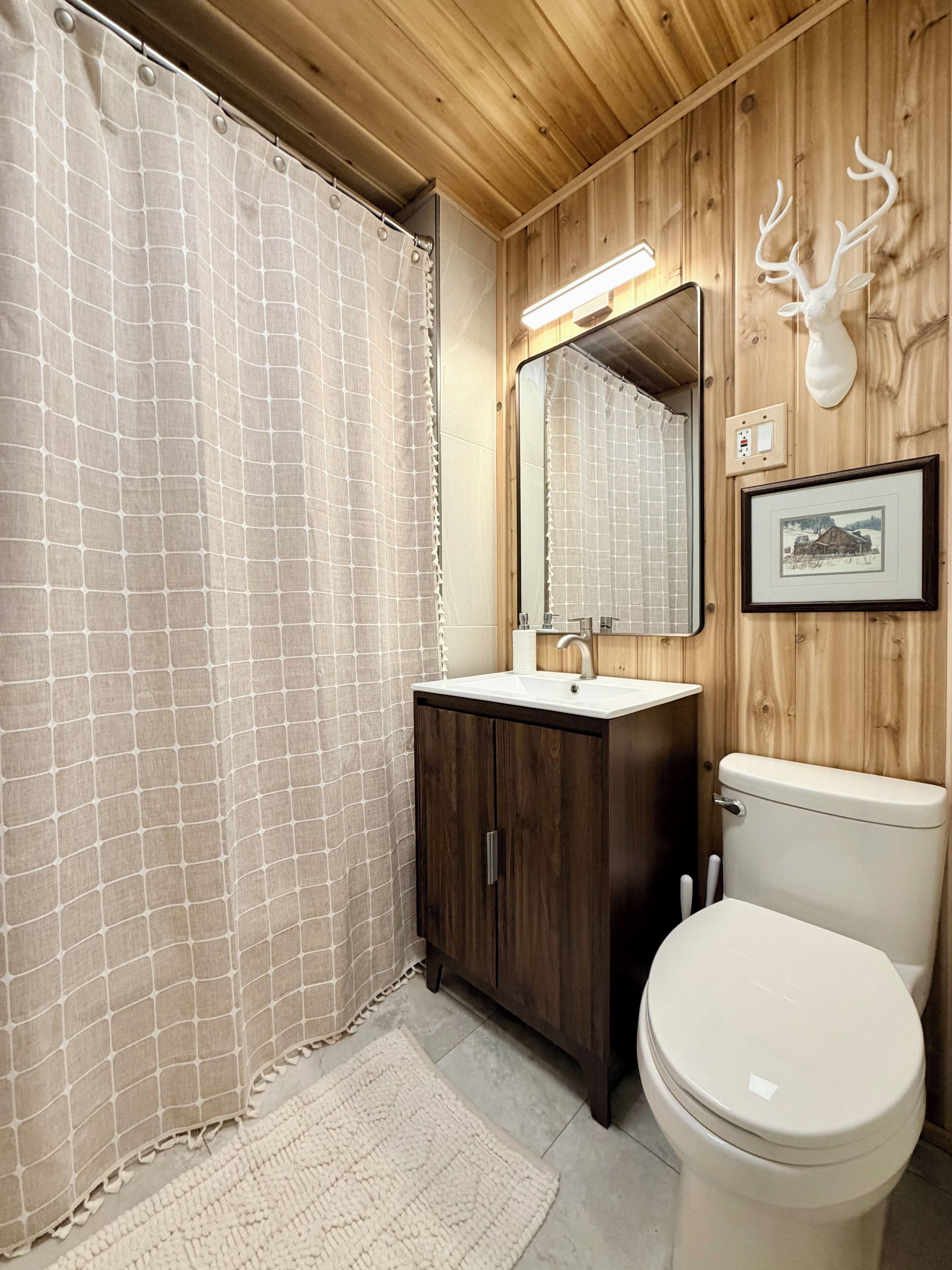 Une salle de bain avec un mur en bois, des toilettes blanches, un lavabo avec un miroir et un tableau encadré.