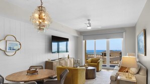 Una sala de estar moderna con una mesa redonda de madera, sillas de mimbre, un sofá amarillo y un sillón beige. Un televisor de pantalla plana de gran tamaño está montado en la pared, y se ve un ventilador de techo. La habitación tiene ventanas grandes que ofrecen vistas a la playa y al océano.