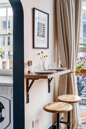 Une cuisine avec une fenêtre, un tableau encadré, une petite table avec des verres et des tabourets.