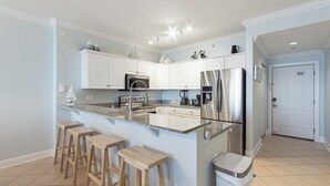 A modern kitchen with white cabinets, a stainless steel refrigerator, a granite countertop, and wooden stools. - Emerald Isle 1102 - Front row beach chair service included! Affordable rates! (Panama City Beach)