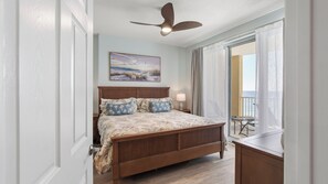 Une chambre avec un lit en bois, un ventilateur de plafond, un tableau et une vue sur l’océan depuis un balcon.