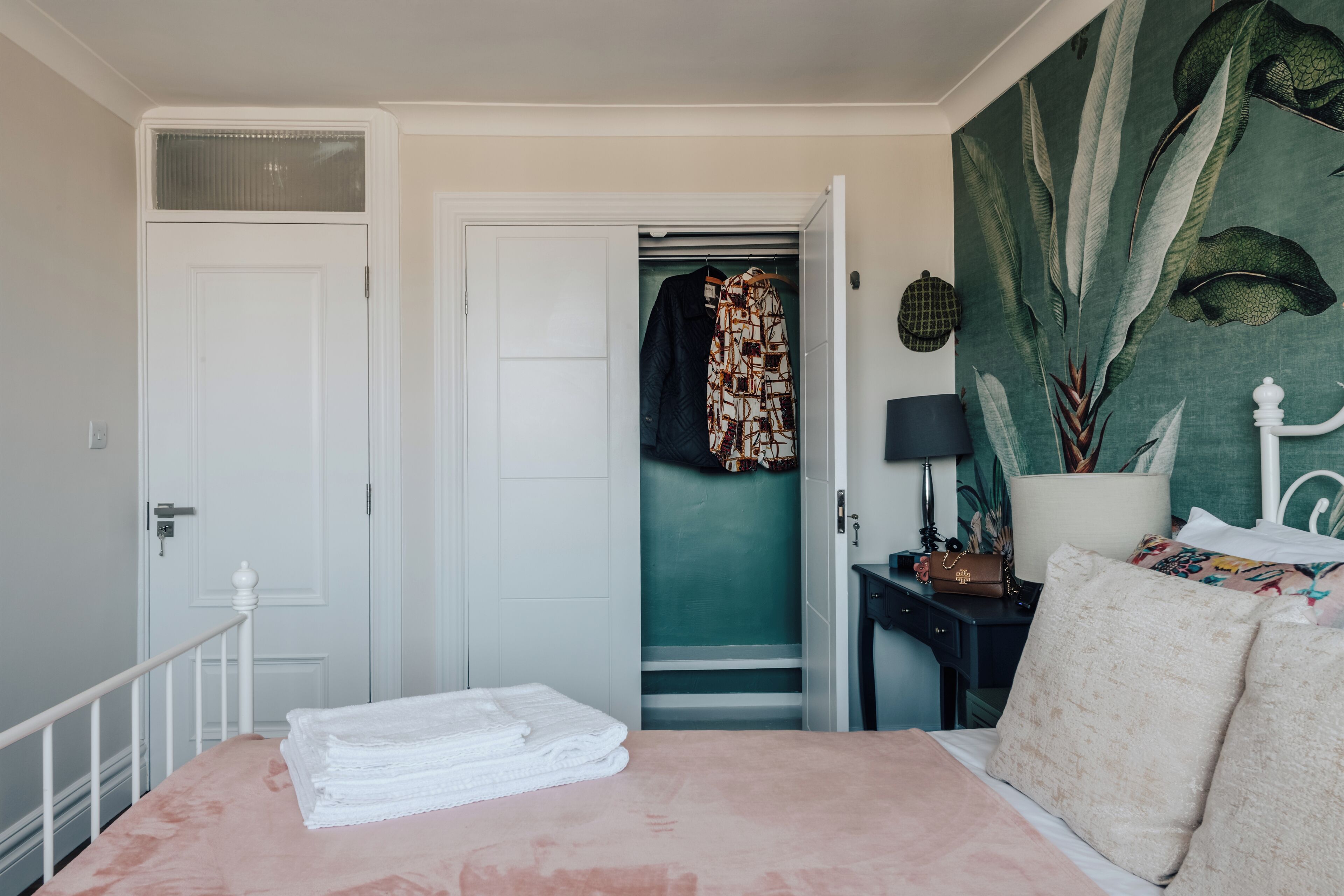 A bedroom with a bed, a closet, a nightstand, and a wall with a large plant.