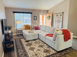 A living room with a sectional sofa, a patterned rug, a small side table, and a door leading to another room.