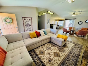 A living room with a sectional sofa, ottoman, patterned rug, and a dining area with chairs and a table. - The Desert Rose | Ground-Level, Garage, 3BR + $510 in complimentary daily activities (Mesquite)