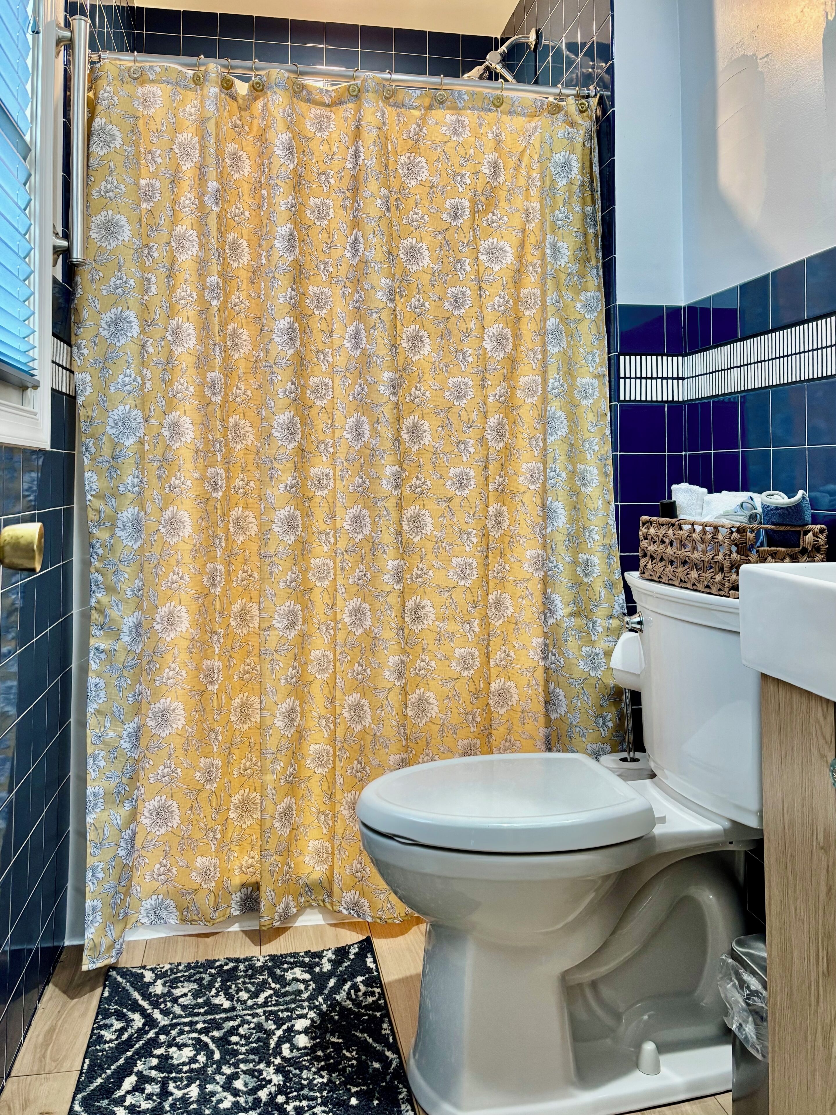 Ein Badezimmer mit gelbem, floralem Duschvorhang, einer weißen Toilette, einem schwarz-weißen Teppich und einer blauen Fliesenwand.
