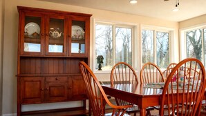 A dining room with a wooden table and chairs, a cabinet with glass doors, and large windows with a view of trees. - Private Sauna, Fireplace and Country Views (Mount Airy)