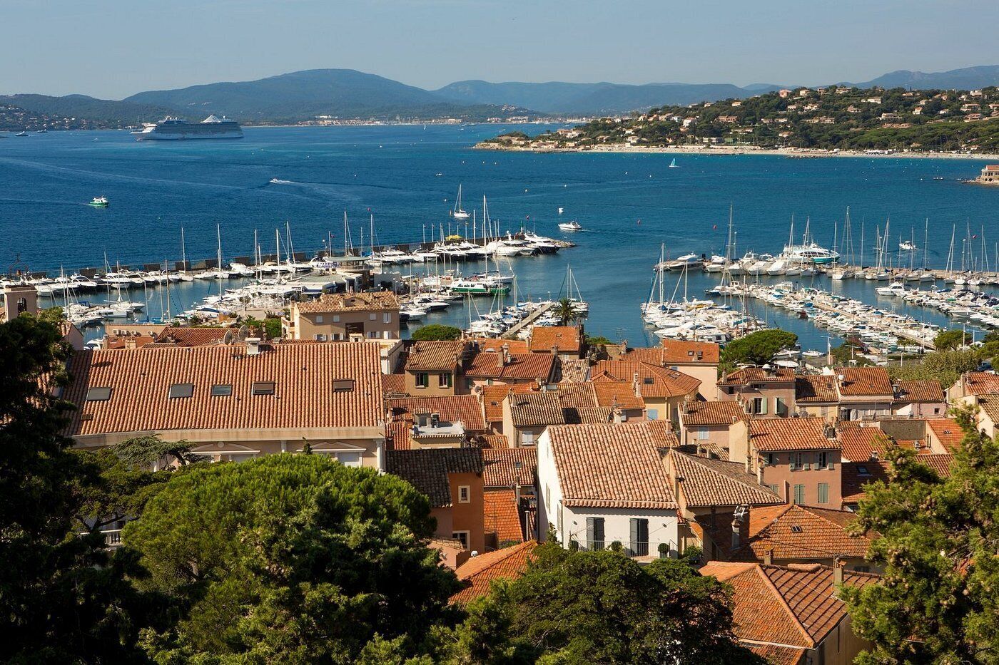 Une ville côtière dotée d’un port de plaisance rempli de bateaux, de bâtiments aux toits de tuiles rouges et offrant une vue dégagée sur la mer et les collines lointaines.
