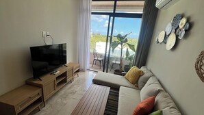 En moderne stue med et fladskærms-TV, træmøbler, en sofa med farverige puder og en balkon med udsigt over grønne områder.