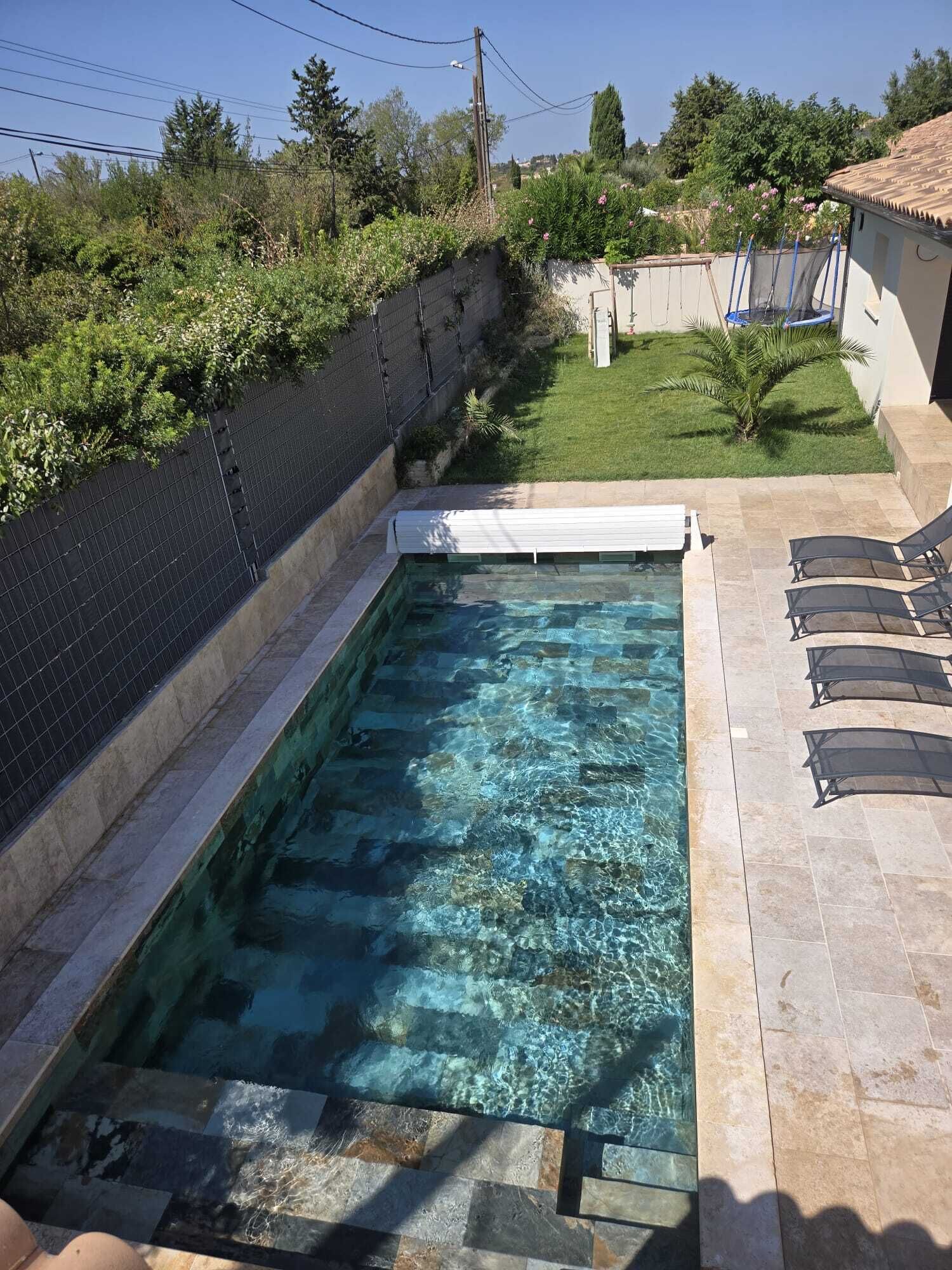 A rectangular swimming pool with clear water, surrounded by a tiled deck and a small grassy area with a fence.