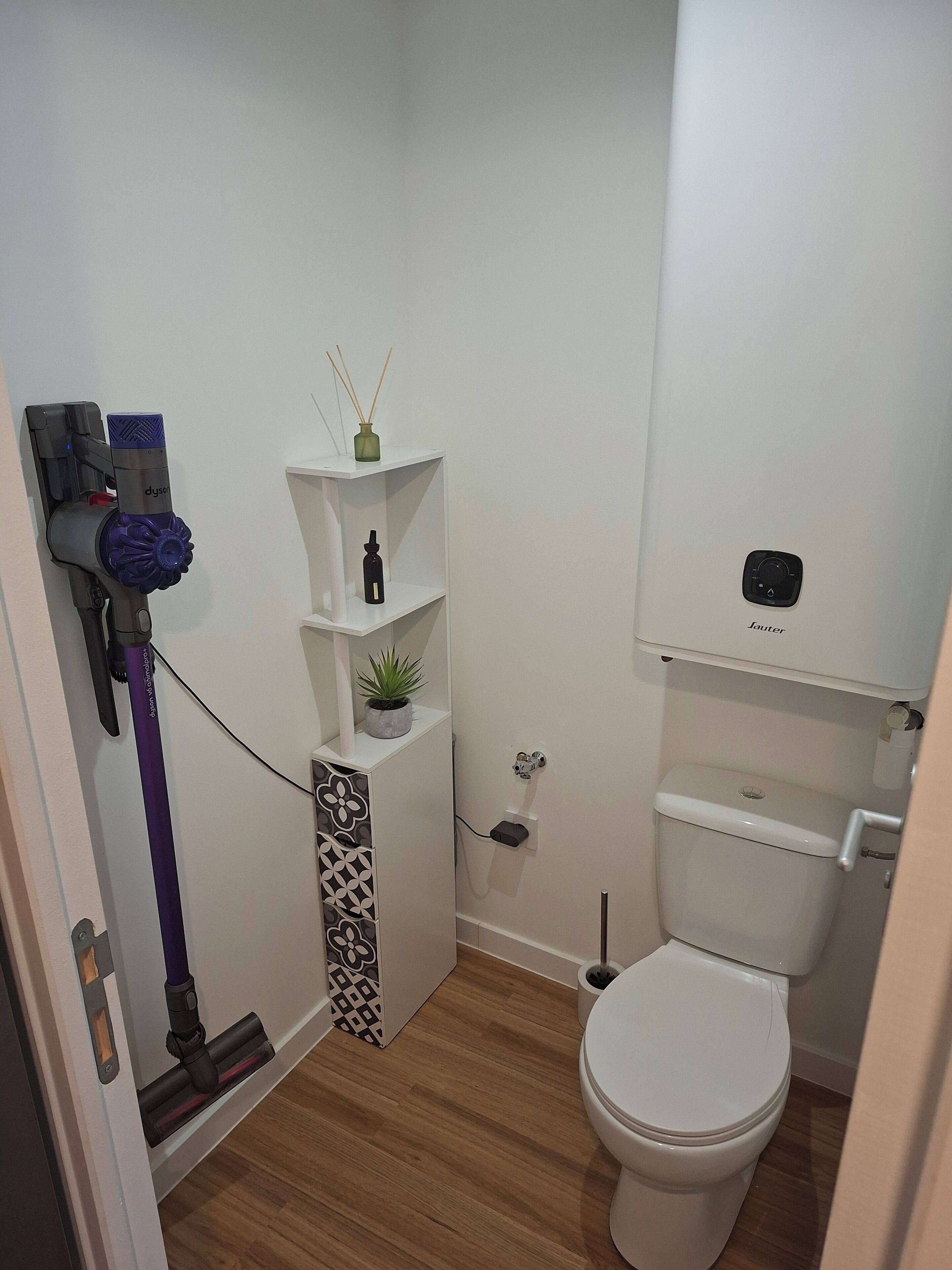 Une petite salle de bain avec des toilettes blanches, une petite étagère ornée d’une plante, un aspirateur Dyson et un porte-rouleau de papier toilette.