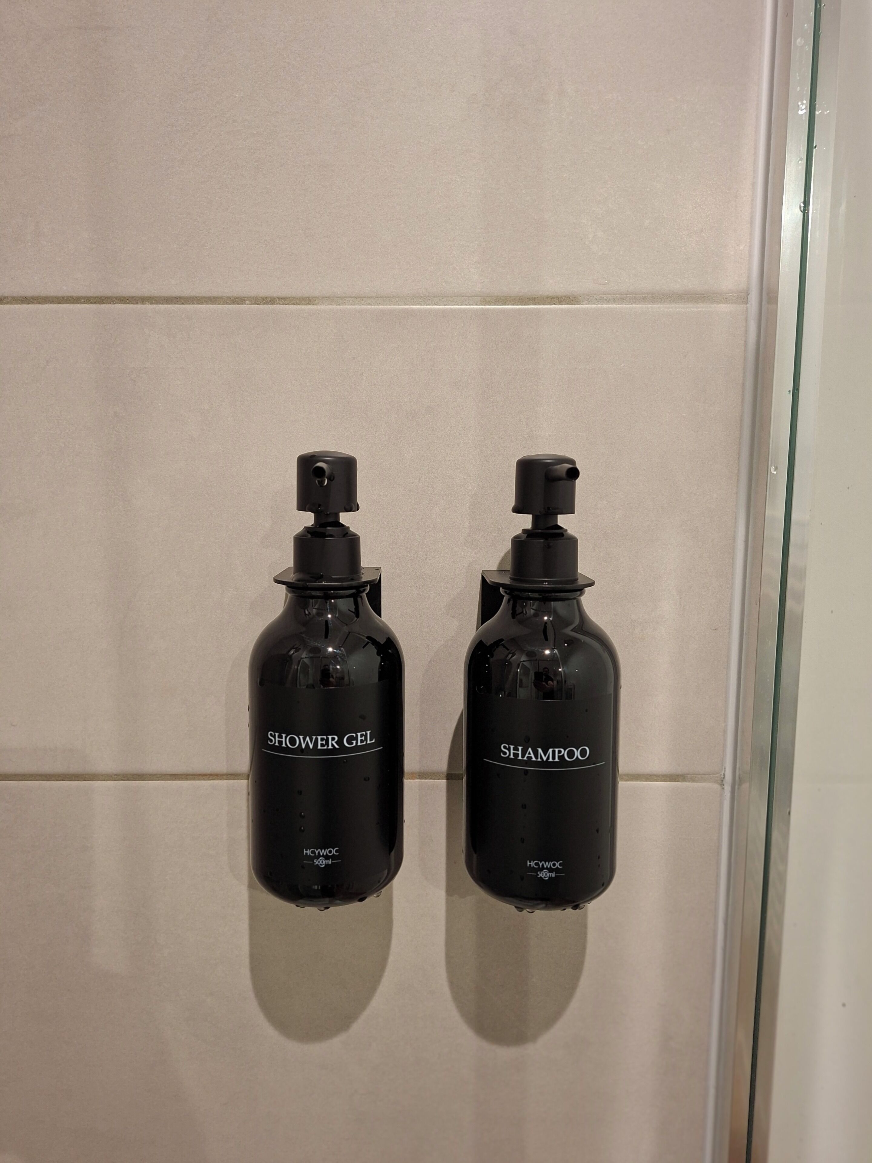 Two black bottles labeled "SHOWER GEL" and "SHAMPOO" mounted on a tiled wall.