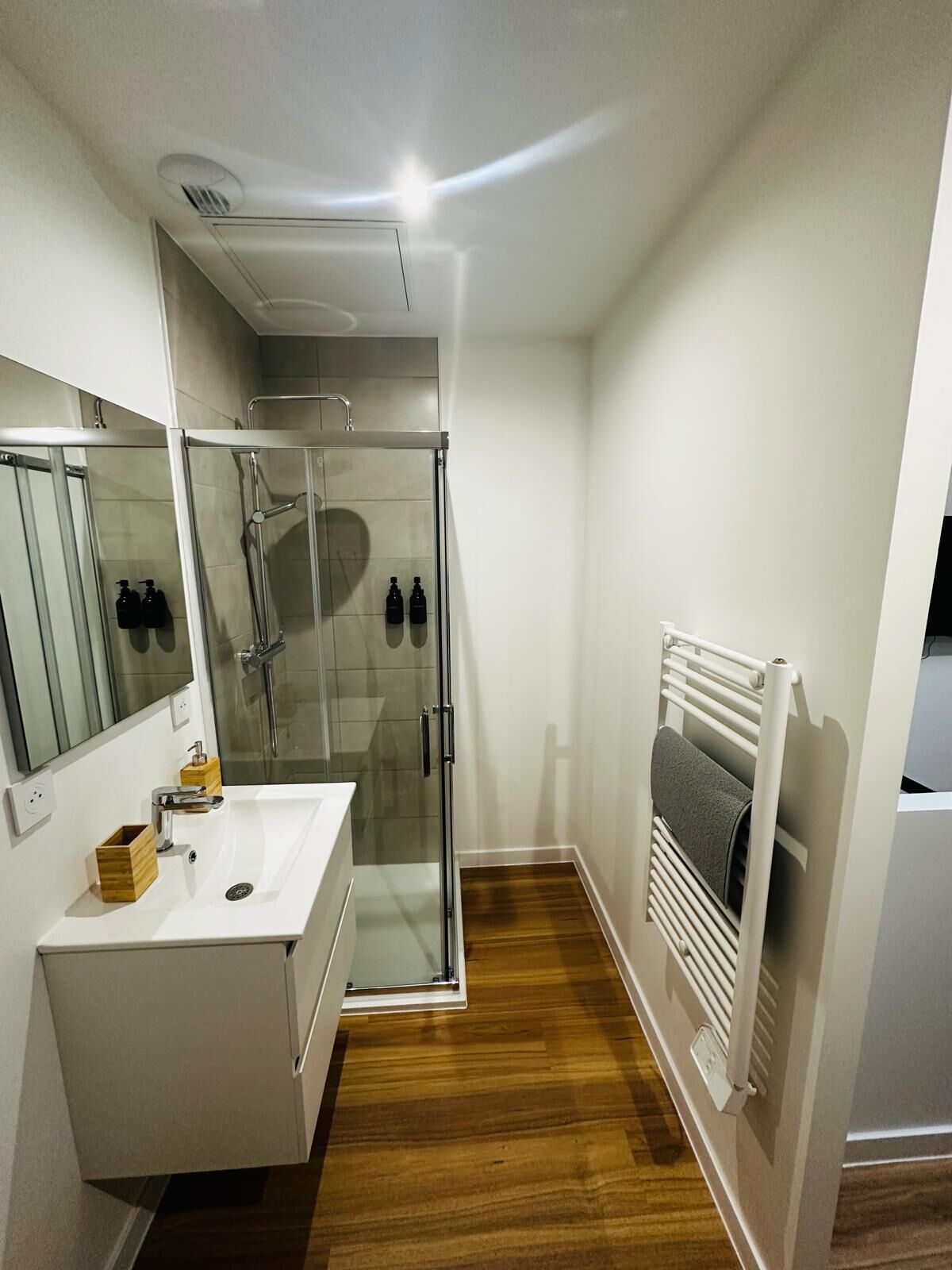 Un baño moderno con cabina de ducha de vidrio, un lavamanos blanco con un soporte de madera para jabón y un perchero con una toalla.