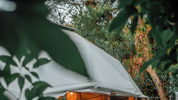 A tented accommodation with a glass door, a bed inside, and a person standing outside holding a glass.
