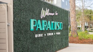 Un mur sur lequel est affiché un panneau indiquant : « Bienvenue à PARADISO » et mentionnant : « REPAS + BAIGNADE + AMBIANCE ».