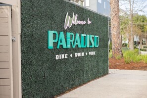 A wall with a sign that reads 'Welcome to PARADISO' and mentions 'DINE + SWIM + VIBE'. - 2BR Home | Grill + Free Parking | Near Beach + DT (North Charleston)