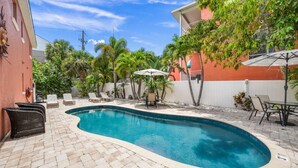 A swimming pool with lounge chairs and umbrellas, surrounded by palm trees and a red building. - Salty Summer – Steps to Siesta Key Village & Beach (Siesta Key)