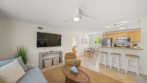 A modern living room with a sofa, coffee table, and a TV. There is a kitchen area with wooden cabinets and a dining table. - Salty Summer – Steps to Siesta Key Village & Beach (Siesta Key)