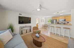 Una sala moderna con sofá, mesa de centro y televisor. Se observa una zona de cocina con armarios de madera y una mesa de comedor.