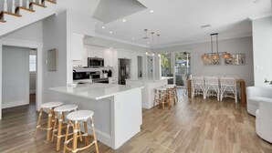 A modern kitchen with white cabinetry, a central island with stools, and a dining area with white chairs. - The Pool House~Steps to Beach & Siesta Key Village (Siesta Key)