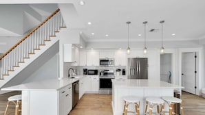 A modern kitchen with white cabinetry, stainless steel appliances, a central island with stools, and a wooden staircase. - The Pool House~Steps to Beach & Siesta Key Village (Siesta Key)