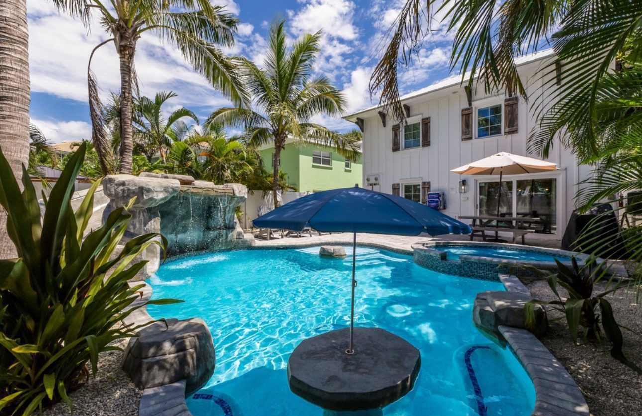 A swimming pool with a waterfall feature, surrounded by palm trees and a building with a green facade.