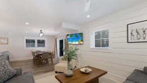 A modern living room with a wooden coffee table, gray sofas, a dining area with a table and chairs, and a large TV screen displaying a beach scene. - Island House – Walk to Siesta Key Beach! (Siesta Key)