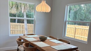 Comedor con mesa de madera, seis sillas, una lámpara colgante y dos ventanas que dan a una valla de madera y árboles.