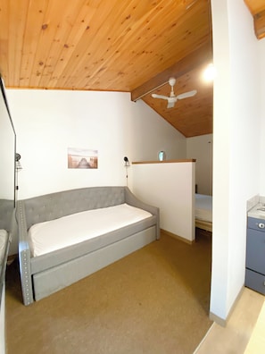 A small, single-bed room with a wooden ceiling and a ceiling fan. There is a small desk with a chair and a picture on the wall.