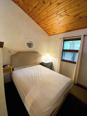 Ein Schlafzimmer mit Holzboden, einem Bett, einem Nachttisch mit Lampe, einem Fenster mit Vorhängen und einem kleinen runden Spiegel an der Wand.