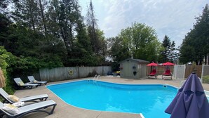 Une piscine rectangulaire bordée de blanc, entourée d’une terrasse en béton. On y trouve des chaises longues et un petit abri doté d’une cheminée.