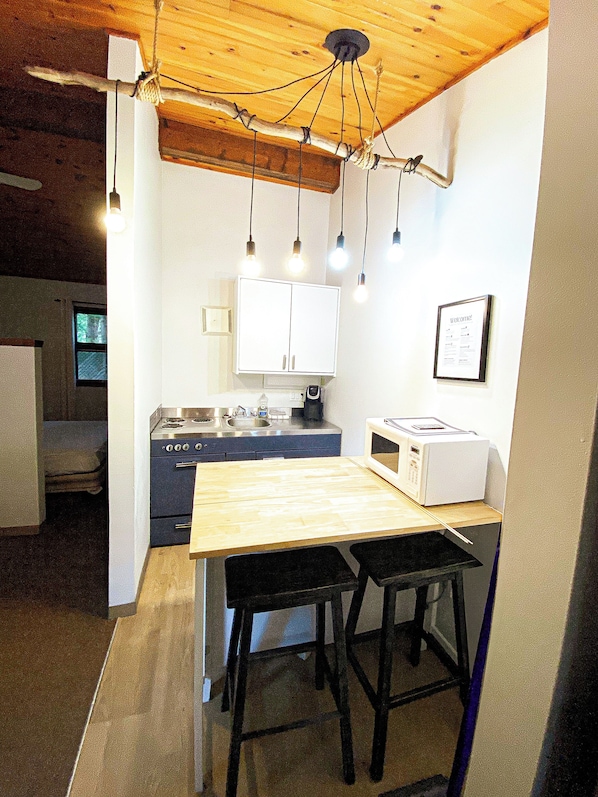 A compact kitchen with a wooden table, two stools, a stove, and a microwave.