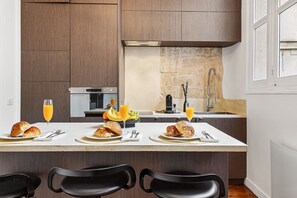 Une cuisine moderne avec des meubles en bois, un pare-feu en pierre et un coin repas où l’on peut prendre le petit-déjeuner avec des croissants et du jus d’orange.