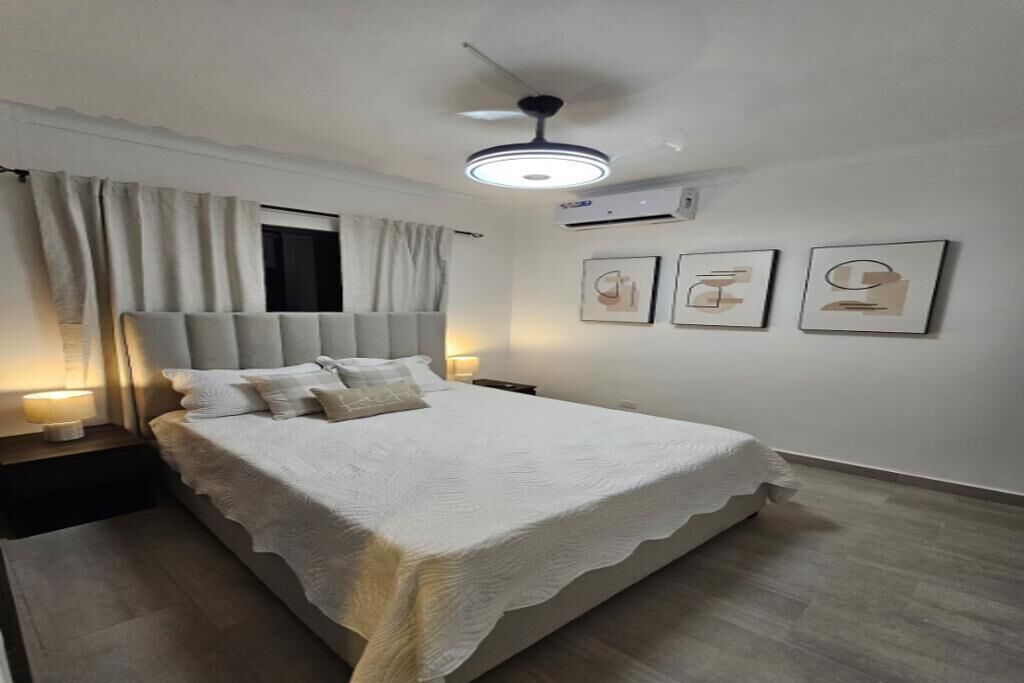 A bedroom with a bed, nightstands, a ceiling fan, and wall-mounted air conditioning unit.