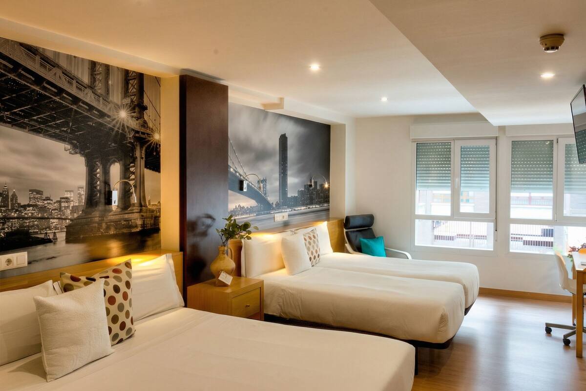Habitación de hotel con dos camas, un escritorio, una silla, un cuadro de gran formato con un paisaje urbano y una ventana con persianas.
