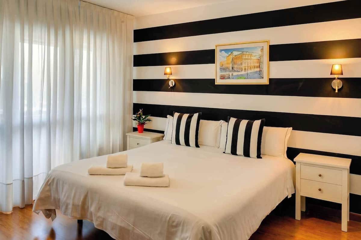 Una habitación de hotel con una cama grande, cabecera a rayas blancas y negras, dos lámparas de noche, una mesita de noche y una ventana con cortinas translúcidas.
