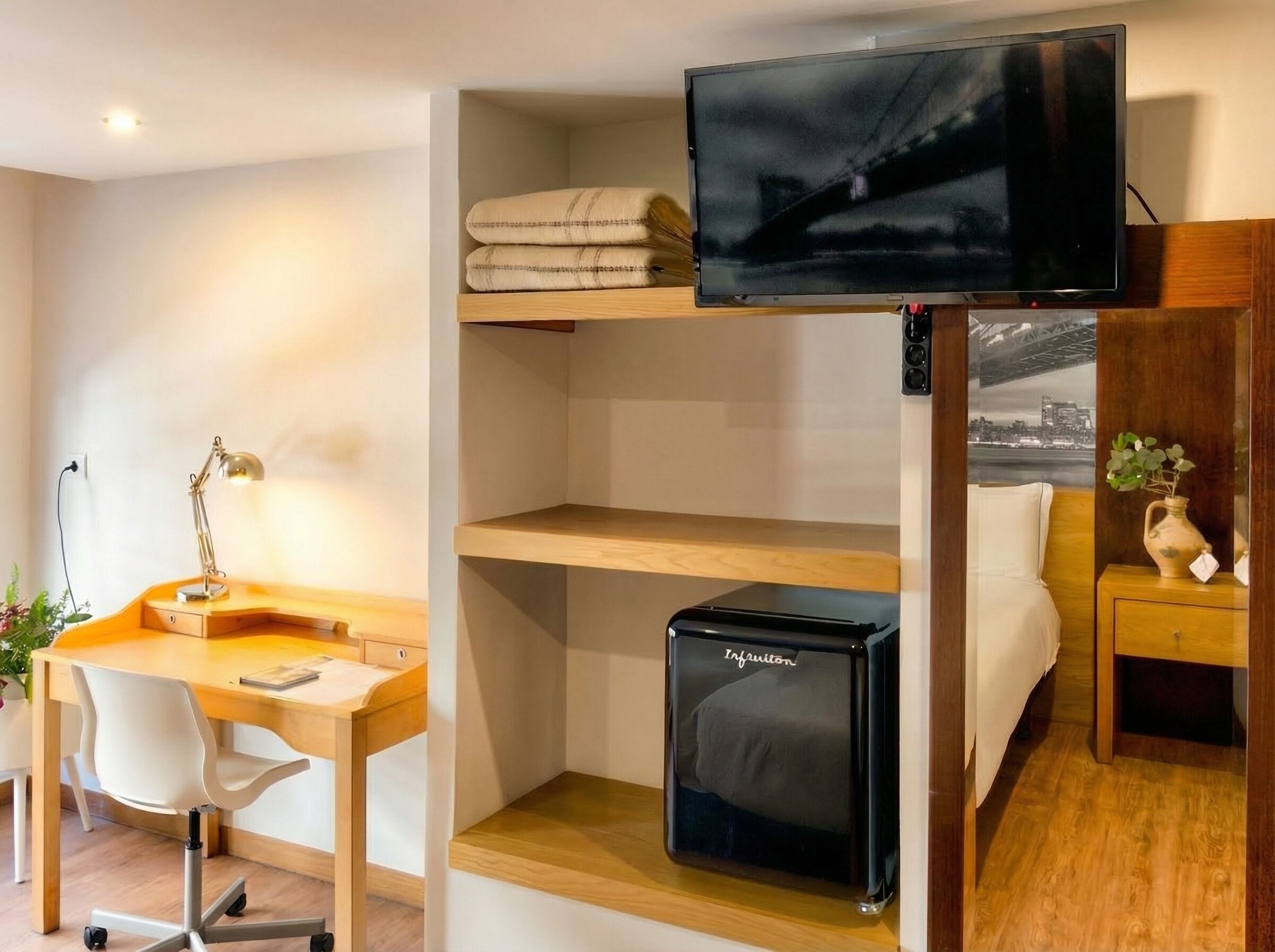 A hotel room with a desk, chair, bed, TV, storage unit, and a view of a cityscape.