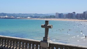 A coastal scene with a large cross monument in the foreground, a sandy beach, and a cityscape in the background. - Hospedaje Boutique Weber's (Gijón)