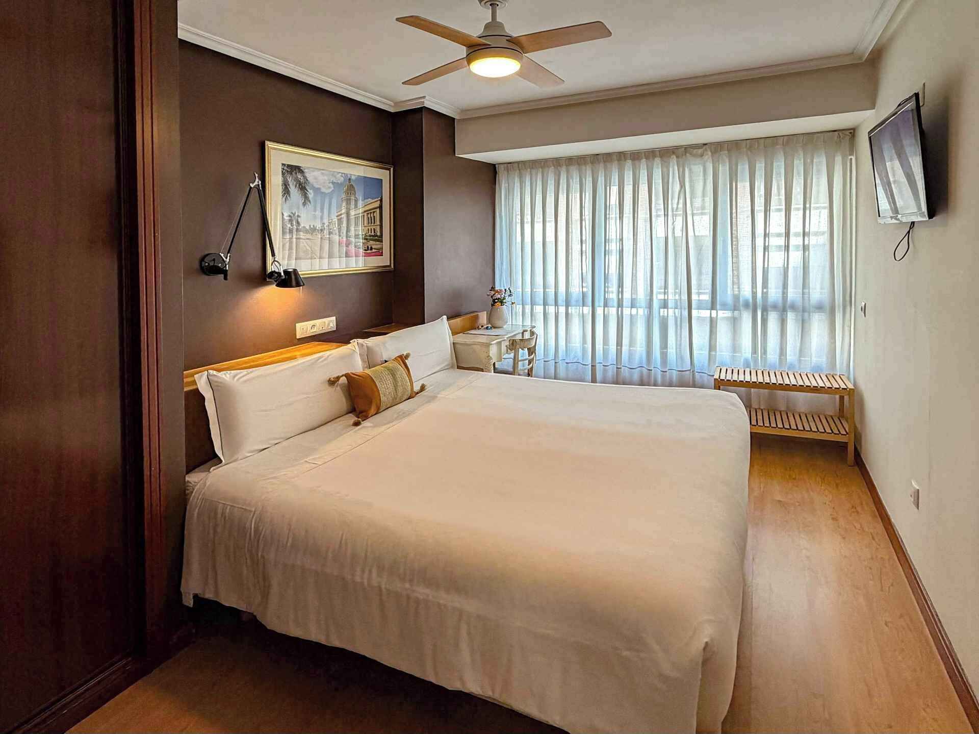 Quarto de hotel com uma cama grande, ventilador de teto, TV fixada na parede e uma janela com cortinas finas.