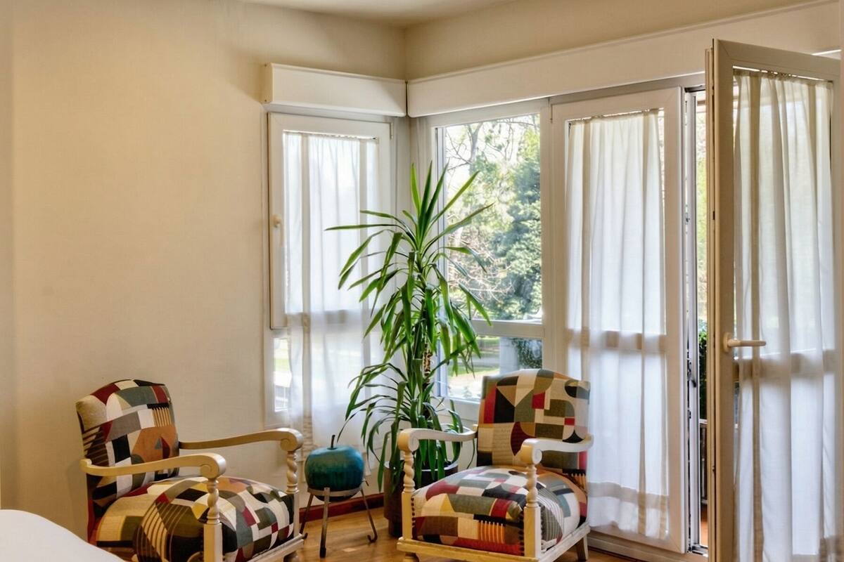 Un dormitorio con una cama, dos sillas, una planta en maceta y un ventanal corredizo que da a un balcón.