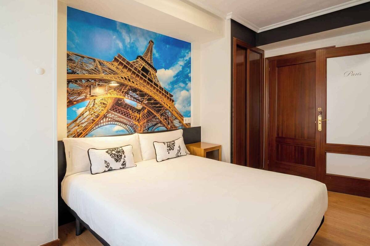 Un dormitorio con un gran mural en la pared de la Torre Eiffel, una puerta de madera y una cama con ropa de cama blanca.