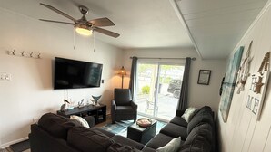 A living room with a dark-colored sofa, a flat-screen TV, a ceiling fan, and a window with a view of the outside. - Cozy Little Slice of OCMD Heaven (Ocean City)
