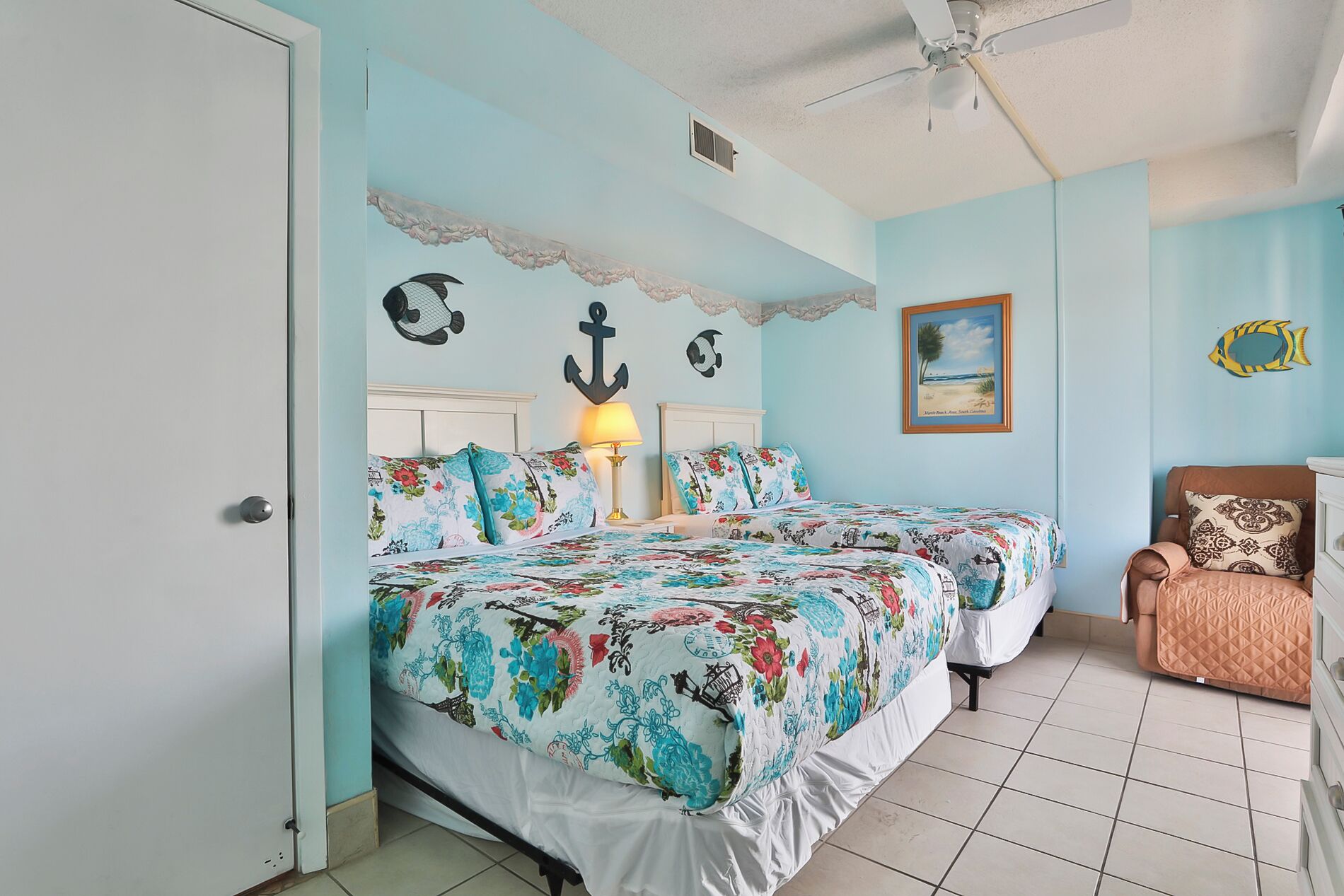 A bedroom with a bed, a nightstand, a lamp, a chair, and wall decorations including an anchor and a fish.