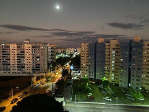 Une vue nocturne d’une ville, avec de hauts immeubles, une rue bien éclairée et une lune visible.