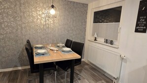 A dining area with a wooden table set for four, black chairs, a hanging light fixture, and a window with blinds.