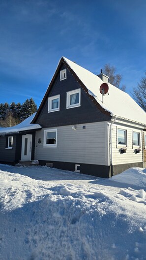 Una casa de dos pisos con techo oscuro y revestimiento blanco, rodeada de nieve.