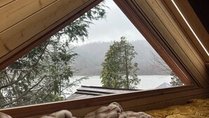 Una camera da letto accogliente in mansarda, con soffitto spiovente, travi in legno e vista su lago e bosco attraverso una finestra triangolare.