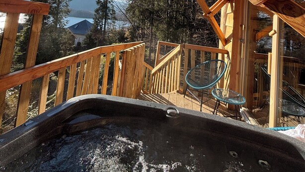 Una terrazza in legno con una vasca idromassaggio, ringhiere in legno e vista su una montagna e un lago.