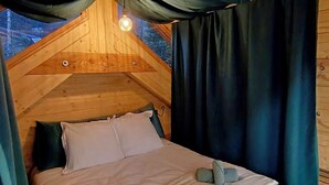 Una stanza a forma di tenda triangolare e accogliente, in legno, con un letto, cuscini e una lampada a sospensione.